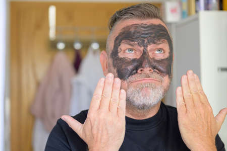 Pleased smiling man using a cosmetic face mask gesturing with his hands towards his face in a skin care and anti-ageing conceptの写真素材