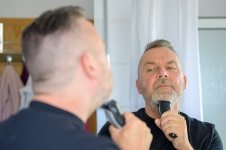 Middle-aged man trimming his beard in the mirror in an over the shoulder view to his face and hand...