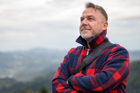 Middle aged grey-haired man with a beard and a lumberjack jacket looks into the distance on a mountain in the fogの写真素材