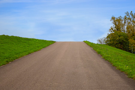 Completely empty spuky road uphill, green grass on the sides and the blue sky behind