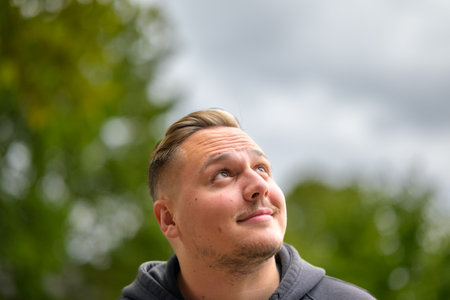 Young man with a mustache and unshaven looks up at the sky with an expectant look and a slight smile in a park or gardenの写真素材