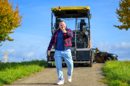 Young man with a commanding smile wearing a red and blue lumberjack jacket and blue t-shirt points to the camera and walks towards the camera with agricultural machinery in the backgroundの写真素材