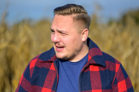 Sideview of an young man with a red and blue lumberjack jacket and blue t-shirt in front of an autumn corn field as a backgroundの写真素材