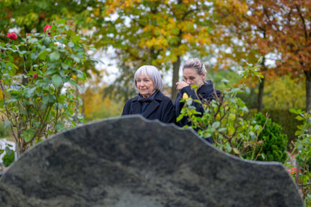 Close up of mother and daughter standing at the man's grave in the cemetery, the mother looking into the distance, the daughter wiping her nose, on a cloudy cold dayの写真素材