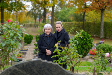 Mother and daughter standing at the man's grave in the cemetery on a cloudy cold day and looking downの写真素材
