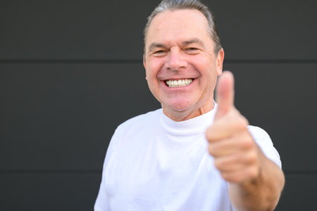A smiling mature man showing his new teeth and giving a thumbs up gesture in front of a black wallの写真素材