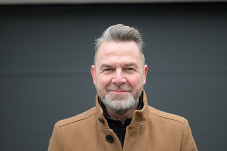 Portrait of a middle-aged man with gray hair and a gray beard in front of a dark wall looking friendly to the cameraの写真素材
