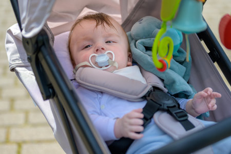 A baby with a pacifier relaxes in a stroller, holding a soft toy and enjoying the outdoors.