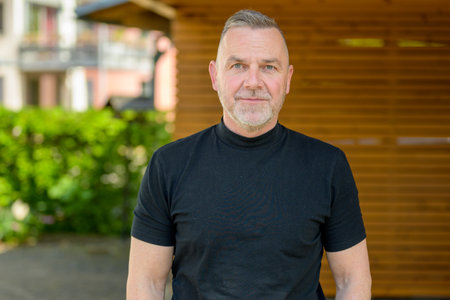 A mature man with gray hair and a beard looks seriously at the camera. Wearing a black turtleneck, he stands against a wooden background with greenery in the blurred distanceの写真素材