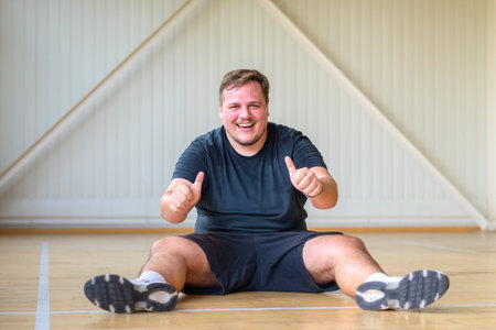 A cheerful man sits on a wooden gym floor, wearing a black t-shirt and shorts, smiling and giving two thumbs up.の写真素材