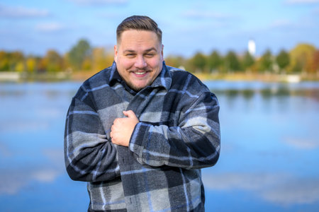 A man stands by a tranquil lake on a brisk autumn day, wrapped in a cozy plaid coat.の写真素材