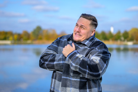 A man stands by a serene lake on a cold day, wrapped in a large plaid coat, visibly feeling the chill.の写真素材
