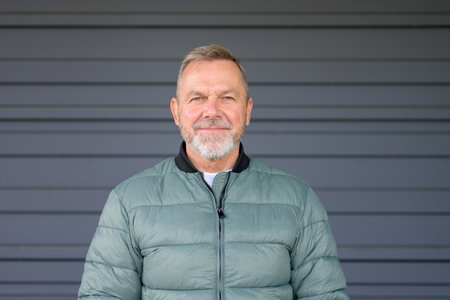 A mature man with a gray beard and a composed expression stands in front of a plain background, wearing a light green puffer jacket.の写真素材