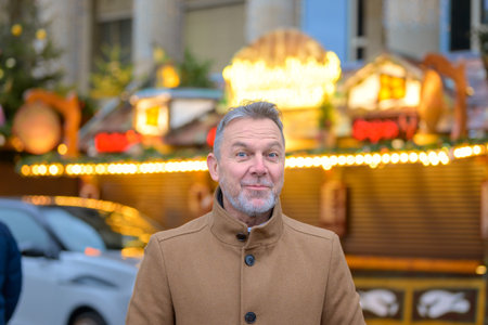 A cheerful man in a beige coat flashes a playful expression against the backdrop of a brightly lit Christmas market stallの写真素材