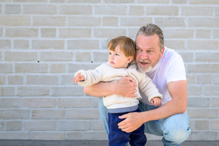 A grandfather crouches beside his toddler in front of a light brick wall, gently holding the child while both gaze intently in the same direction.の写真素材