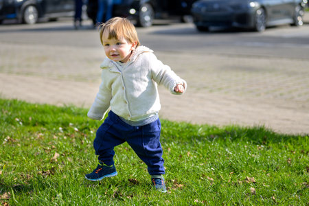 A joyful toddler walks confidently through a grassy area, dressed in a cozy hoodie and blue pants.