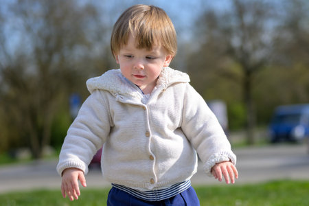 A toddler in a cozy beige hoodie and blue pants stands outdoors on a sunny day, gazing down with a...