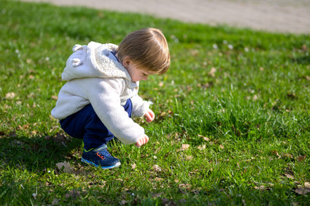 A young child squats intently in a sunlit park, closely examining the grass and small leaves.