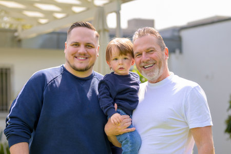 Three generations of one family stand together in a sunlit garden â€“ grandfather, father, and...