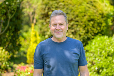 A senior man stands confidently in a lush green garden, looking directly at the camera with a gentle...