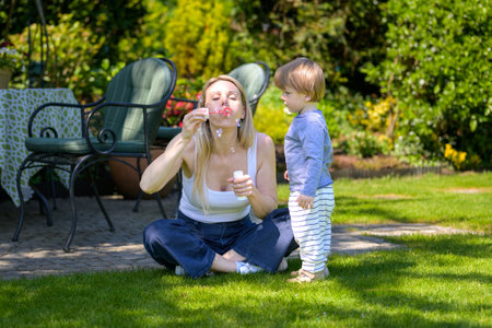 A young mother blows soap bubbles while her toddler son stands nearby, watching with curiosity.