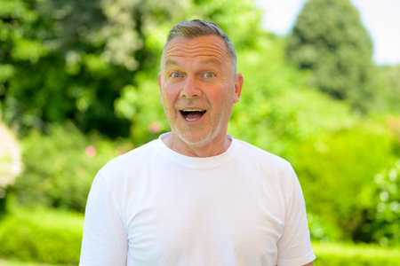 Cheerful senior man in a white t-shirt expressing surprise with wide open eyes and mouth, standing outdoors in a green garden.の写真素材