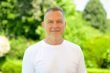 Older man wearing a white t-shirt standing calmly outdoors in a bright, green park setting.の写真素材