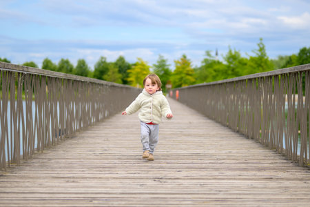Little boy in cozy fleece jacket running joyfully across a long wooden bridge on a cloudy spring day.の写真素材