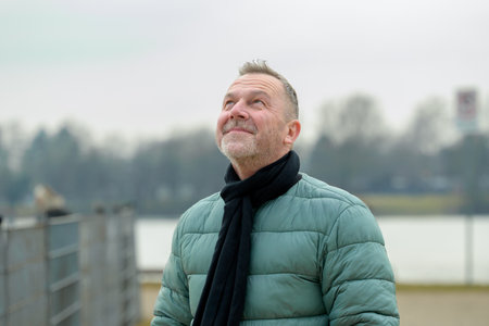 A mature man in a green quilted jacket and black scarf looks upward with a peaceful, thoughtful...
