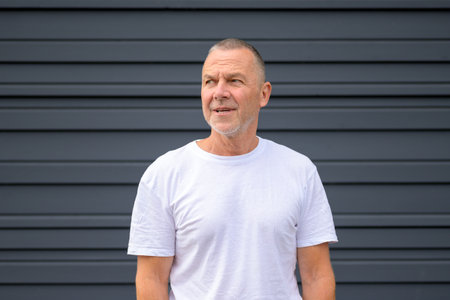Portrait of a smiling senior man wearing a plain white t-shirt, standing in front of a modern dark...