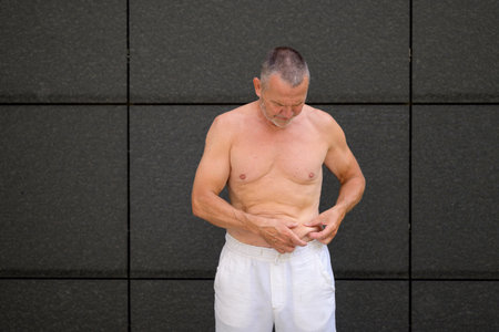 Shirtless senior man standing in front of a dark tiled wall while pinching and inspecting his belly fat.の写真素材