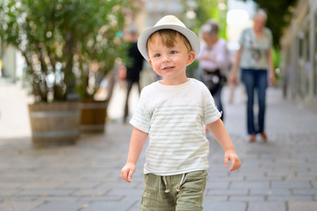 Joyful toddler boy wearing a straw summer hat and striped shirt walking along a sunny city street.の写真素材