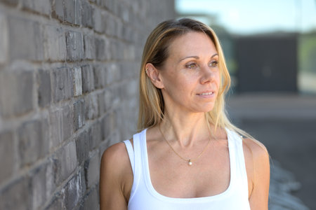 A blonde woman with natural makeup and a gold necklace stands next to a textured brick wall, gazing calmly into the distance.の写真素材