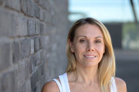 Natural close-up portrait of a smiling blonde woman in a white top, standing outdoors near a gray brick wall.の写真素材