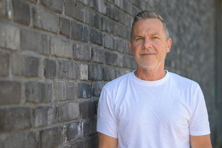 Portrait of a mature man in a white T-shirt leaning against a gray brick wall, facing the camera with a calm and confident expression.の写真素材