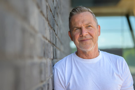 Close-up portrait of a mature man with short gray hair and a white T-shirt, leaning against a brick wall.の写真素材