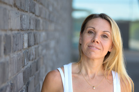 A confident blonde woman in a white tank top gently tilts her head while standing in front of a gray brick wall.の写真素材