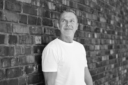Black and white portrait of a confident senior man in a white shirt standing against a textured brick wall.の写真素材