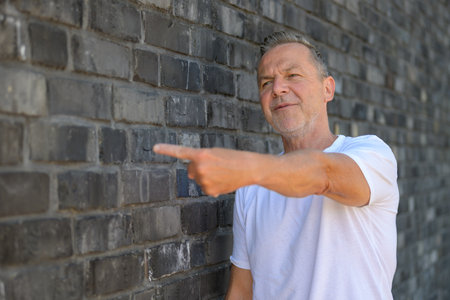 Confident mature man wearing a white T-shirt standing in front of a gray brick wall.の写真素材