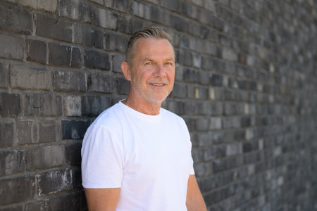 Portrait of a smiling senior man leaning casually against a brick wall.の写真素材