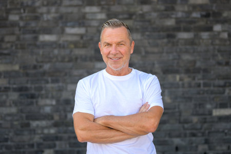 Smiling senior man standing confidently in front of a dark brick wall with his arms crossed.の写真素材