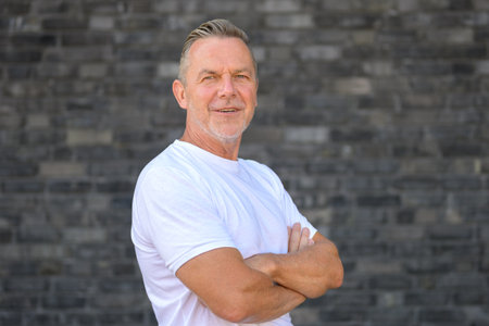 Portrait of a confident senior man standing in front of a dark brick wall with arms crossed.の写真素材