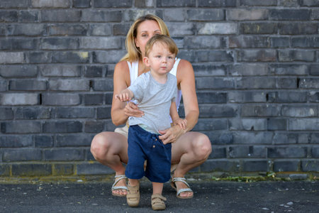 A young woman crouches behind a toddler boy, gently holding his shirt as he takes a step forward.の写真素材