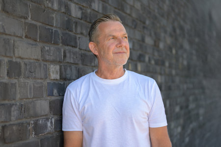 Portrait of a mature man in a white T-shirt leaning against a gray brick wall while looking off into the distance with a thoughtful expression.の写真素材