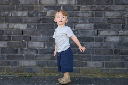 Full-body portrait of a toddler boy standing in front of a gray brick wall, wearing a gray T-shirt, navy blue shorts, and beige shoes.の写真素材