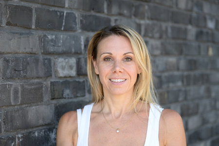 A blonde woman in a white top stands in front of a modern grey brick wall, smiling confidently at the camera.の写真素材