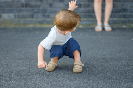 A young toddler squats on asphalt, facing away from the camera, reaching one arm upward toward a standing woman in sandals, whose legs are visible in the background.の写真素材