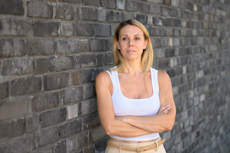 Blonde woman leaning against a textured brick wall, arms crossed, and looking forward with a calm, self-assured expression.の写真素材
