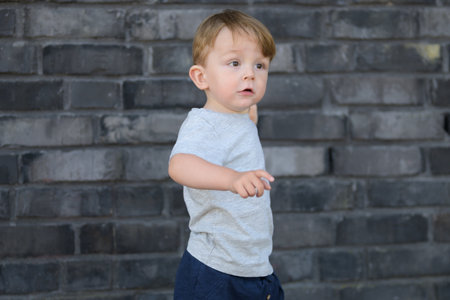Side view of a young toddler boy standing in front of a dark brick wall.の写真素材