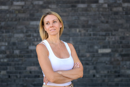 Blonde woman in a white sleeveless top, arms crossed, gazing slightly to the side with a soft, thoughtful expression.の写真素材